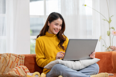 Woman sitting on couch researches how much house can she afford on a laptop.