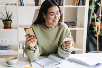 Young woman sits at desk while researching how to pay off credit card debt on her mobile phone.